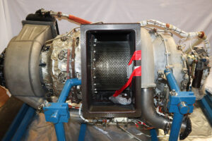 Close-up of an aircraft auxiliary power unit (APU) with exposed interior wiring, metal housing, and visible mechanical components and tubing.